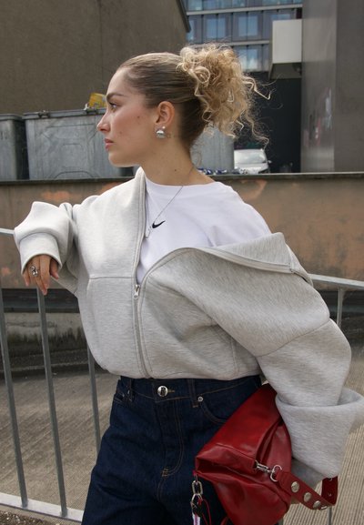 Sudadera gris con cremallera de corte oversize, llevada sobre una camiseta blanca de Nike. Pantalones de mezclilla oscura y un bolso rojo completan el atuendo.