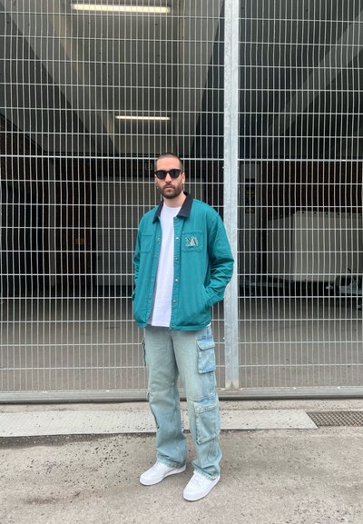 Chaqueta azul verdosa de botonadura con cuello negro, camiseta blanca, pantalones cargo azul claro y zapatillas blancas. Las gafas de sol complementan el atuendo.