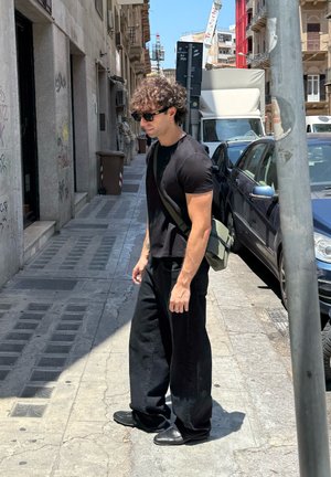 Camiseta de algodón negra, pantalones negros holgados y zapatos negros. Bolsa de hombro verde llevada tipo bandolera. Calle iluminada con edificios y vehículos.
