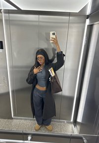 Kvinde iført en lang, mørk frakke over en grå kortærmet top og vidde navy bukser. Hun holder en brun taske og en telefon, omgivet af metalliske elevatorvægge.