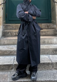 Črn dolg plašč z visokim ovratnikom, zavezan pas in veliki gumbi. Usklajen s črnimi ohlapnimi hlačami in velikimi črnimi športnimi copati. Stopnice v ozadju.