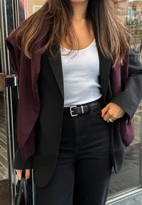 Μαύρο σακάκι φορεμένο πάνω από ένα άσπρο ριγέ tank top, συνδυασμένο με μαύρα παντελόνια ψηλής μέσης και ένα μπορντό πουλόβερ ριγμένο στους ώμους.