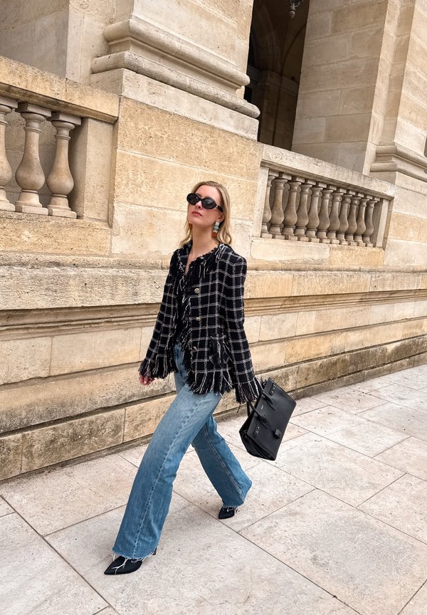 Mujer caminando junto a una balaustrada de piedra, vestida con una chaqueta de flecos a cuadro en blanco y negro, vaqueros azules, gafas de sol negras y llevando un bolso grande negro.