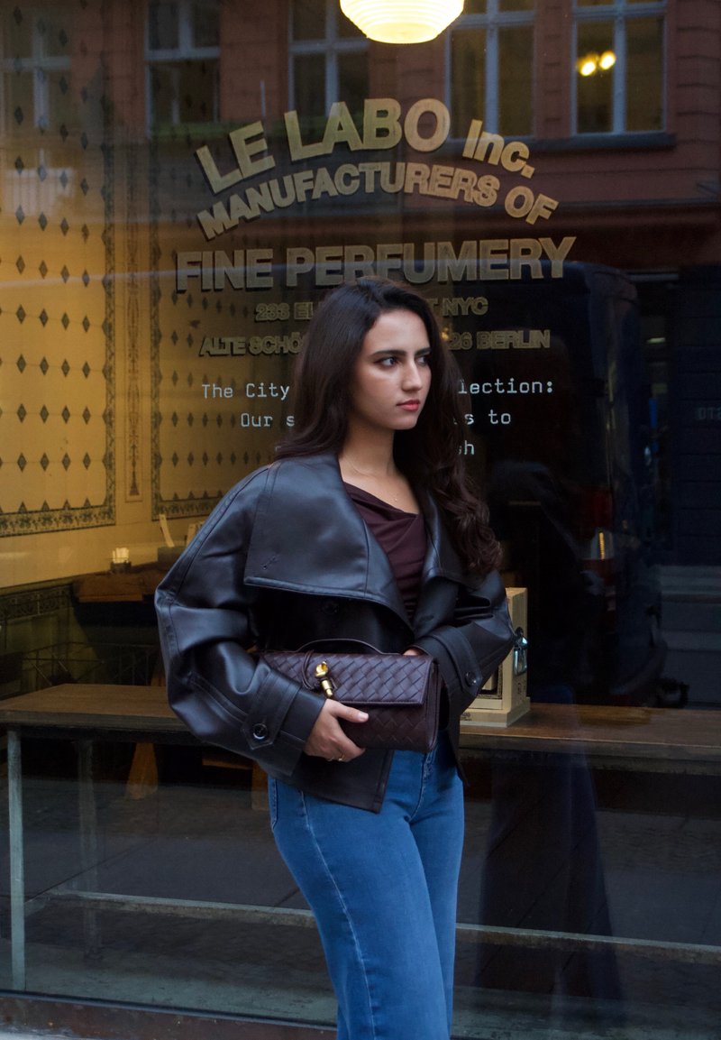 Veste en cuir noir avec un col surdimensionné, haut marron foncé, jeans bleus et un sac marron matelassé avec un fermoir doré, se tenant devant un magasin.