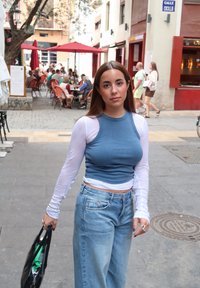 Débardeur côtelé bleu ajusté sous une chemise blanche à manches longues, assorti à un jean large délavé, tenant un sac cabas noir.