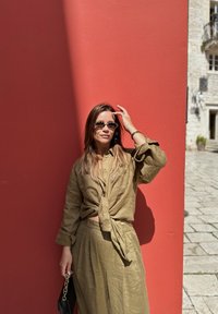 Olivově zelený lněný outfit s uzlovou košilí a volnými kalhotami. Černé sluneční brýle a malá černá kabelka. Červená zeď v pozadí.