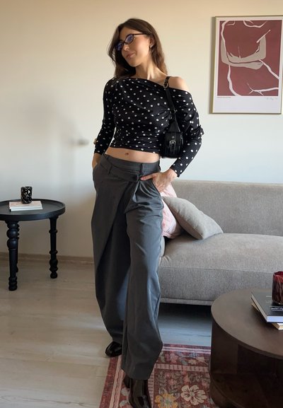 Top negro con hombros descubiertos y lunares blancos, pantalones grises de pierna ancha con pliegues, y botas negras brillantes. Se ve un bolso de mano como acento.