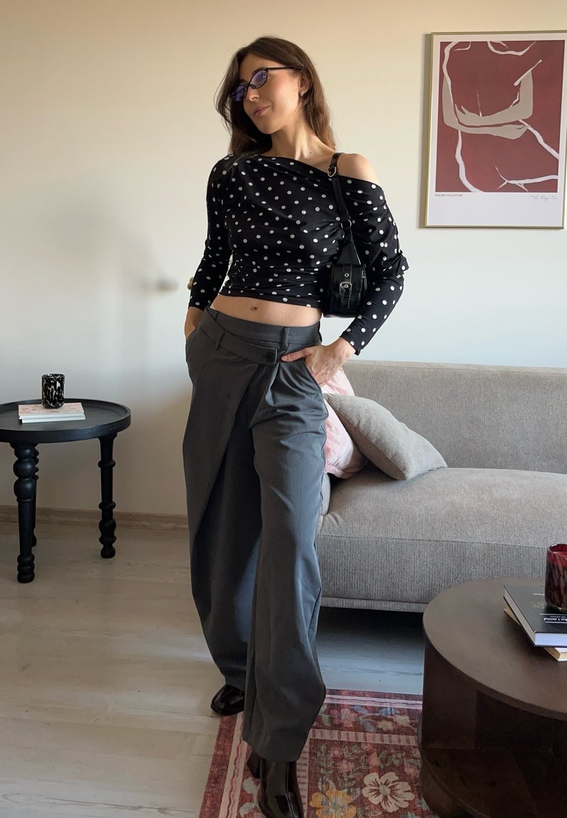 Top nero scollato sulle spalle con pois bianchi, pantaloni grigi a gamba larga con pieghe e stivali neri lucidi. Borsa di accento visibile.