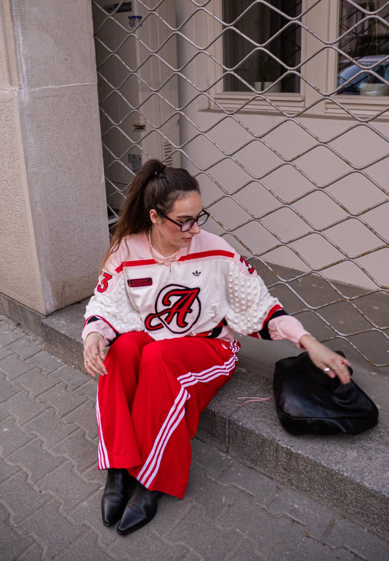 Mladá žena v červených teplákoch Adidas a bielom športovom svetri sedí na obrubníku a siahá na čiernu kabelku vedľa seba.