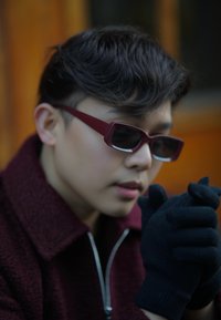 Cappotto bordeaux con zip, guanti in maglia nera e occhiali da sole rettangolari. In primo piano una persona che stringe le mani, con capelli texturizzati.
