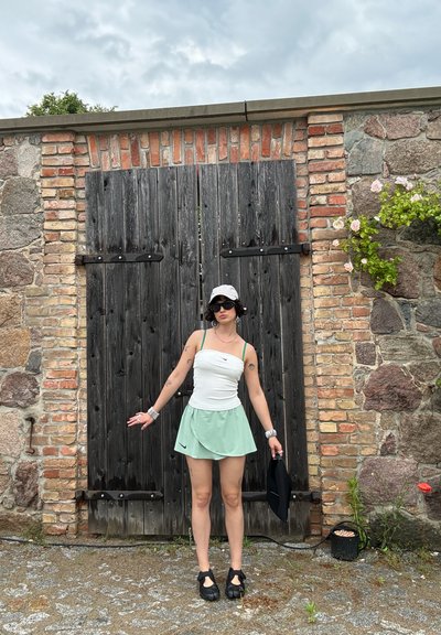 Persona con parte superior blanca, falda mint, sombrero y gafas de sol, está de pie con los brazos ligeramente extendidos frente a grandes puertas de madera negras en una pared de ladrillo y piedra.