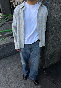 Jasnoniebiesko-biała koszula w kratę na guziki, noszona na białym t-shircie, zestawiona z przetartymi, szerokimi spodniami jeansowymi i czarnymi butami. Tkanina o wyraźnej fakturze.