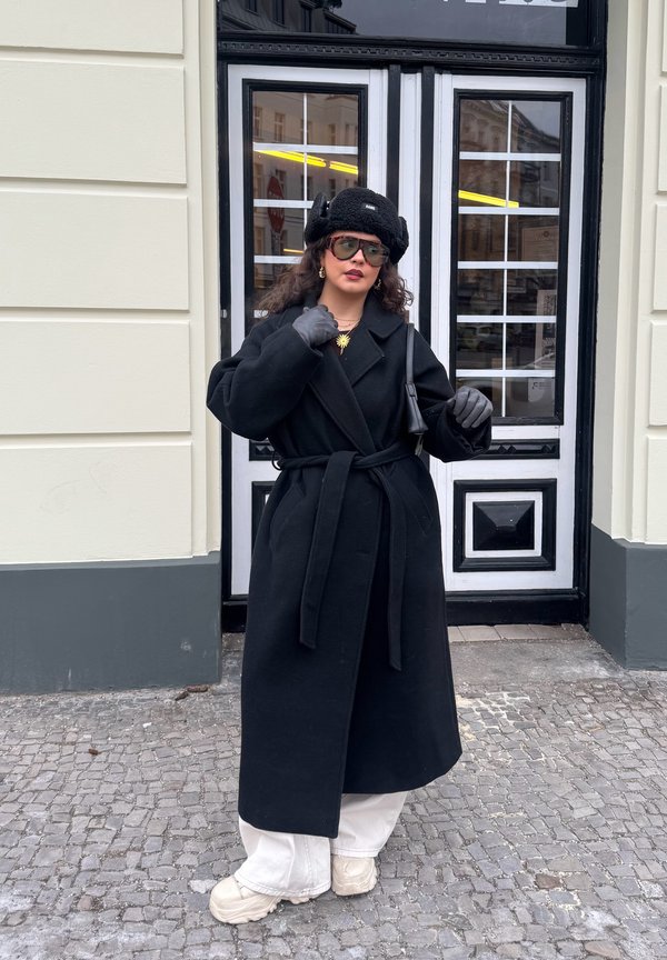 Žena u crnom kaputu, beretki, rukavicama, sunčanim naočalama, bijelim hlačama i debelim cipelama stoji na kaldrmisanoj ulici ispred vrata u crno-bijeloj boji.