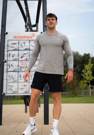 Camisa de manga larga gris y pantalones cortos negros usados por una persona con una estación de fitness de fondo, que presenta instrucciones de ejercicio e íconos.