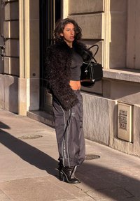 Femme debout sur le trottoir portant une veste noire en matière duveteuse, un top court, un pantalon ample Nike Shox et des bottes noires à talons hauts, tenant un sac à main noir.