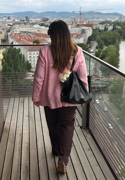 Mujer vistiendo un blazer de lino color rosa claro, pantalones marrón oscuro con rayas blancas, y llevando un bolso negro con flores blancas dentro.