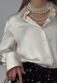 Satin bluzica boje kreme s klasičnom kragnom i dugmadi, složena s više redova perličastih ogrlica, u kombinaciji s crnom suknjom od šljokica.
