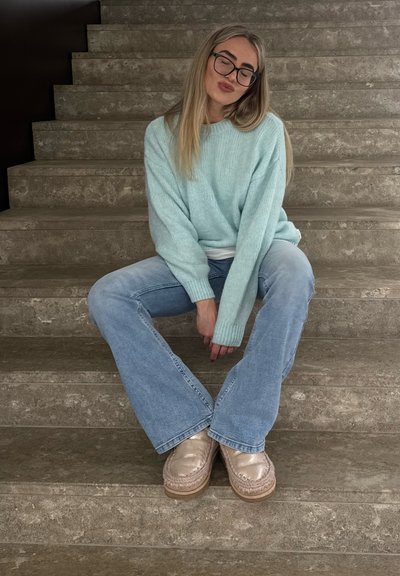 Joven con gafas, suéter azul claro y jeans, sentada en unas escaleras de piedra, con las manos entrelazadas, ojos cerrados y labios fruncidos.