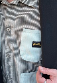 Camicia a righe marroni e bianche con una tasca verde menta dotata di un’etichetta nera. Il tessuto presenta un aspetto strutturato e dettagli con bottoni.