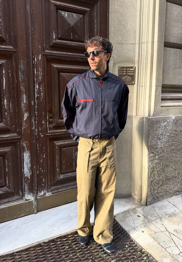 Un hombre con gafas de sol, una chaqueta azul marino y unos pantalones beige está de pie frente a una puerta de madera desgastada.