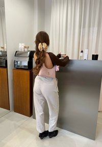 Maglietta a maniche lunghe marrone, borsa rosa con accenti, pantaloni chiari, scarpe nere a punta, postura rilassata al bancone di un caffè.