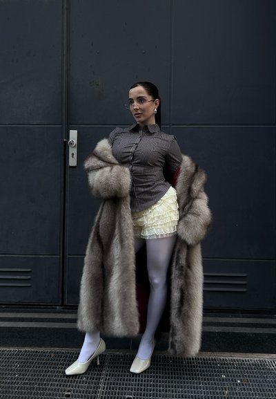 Abrigo de piel sintética en tonos grises con forro rojo, camiseta a rayas, pantalones cortos de encaje y medias blancas. Tacones beige de punta cerrada y gafas redondas.