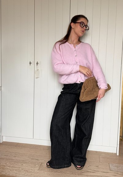 Cárdigan rosa esponjoso con botones, combinado con jeans de pierna ancha negros. Sostenido un bolso de mano texturizado en color marrón. De pie contra un fondo blanco.