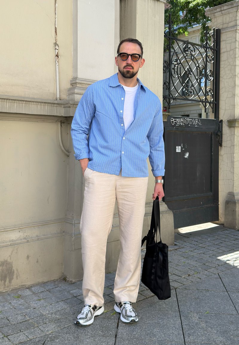 Homme portant des lunettes, chemise à rayures bleues, t-shirt blanc, pantalon beige et baskets, portant un sac noir, debout près d'un bâtiment en pierre et d'une grille en fer forgé.
