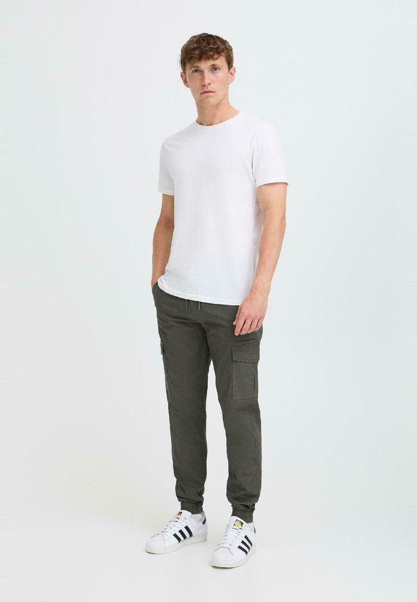 T-shirt bianco a maniche corte, pantaloni cargo verde oliva con tasche e sneakers bianche con righe nere. Outfit semplice e informale.