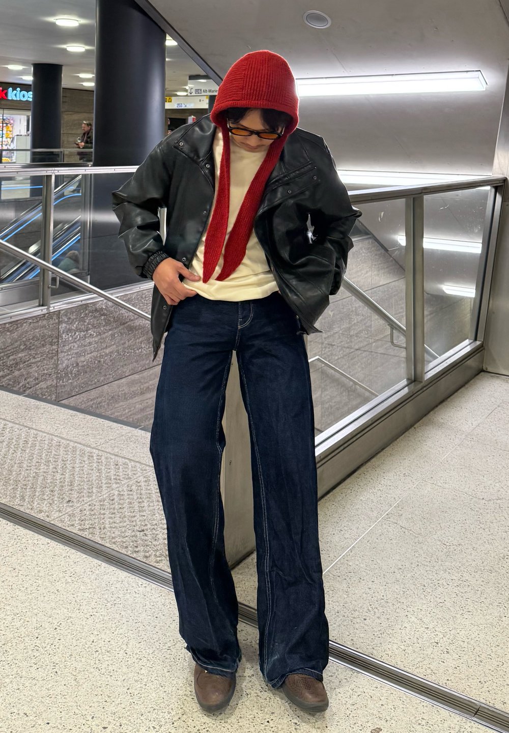 Veste en cuir noire, t-shirt blanc, pantalon en denim bleu long, bonnet tricoté rouge et chaussures marron. Le modèle est posé dans un espace intérieur bien éclairé.