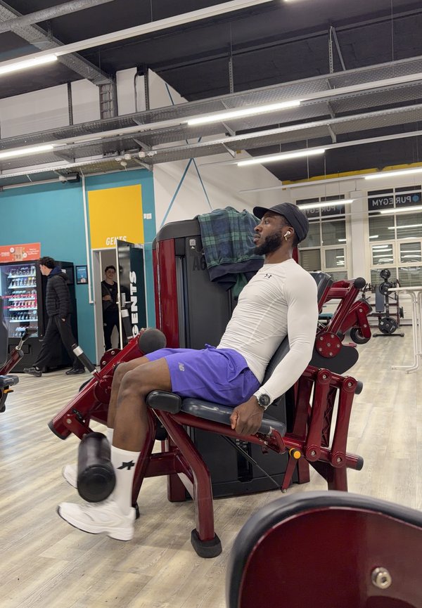 Ein Mann in Sportkleidung benutzt eine Beinstreckermaschine im Fitnessstudio, im Hintergrund sind weitere Personen und Trainingsgeräte zu sehen.