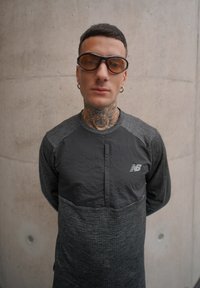 Haut noir à manches longues avec des accents gris texturés, doté d'une fermeture éclair quart, logo New Balance, et modèle portant des lunettes teintées marron.