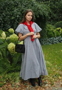 Schwarz-weiße Karomuster-Maxikleid mit Puffärmeln, kombiniert mit einem roten Schal und einer kleinen schwarzen Handtasche, getragen mit schwarzen Stiefeln. Hintergrund mit Grünpflanzen.