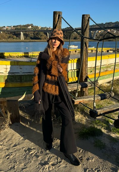 Shawl negro con franjas de piel marrón, guantes de punto y gorro de estampado de leopardo. Pantalones acampanados negros y botas de tobillo negras. Barcos de madera en el fondo.