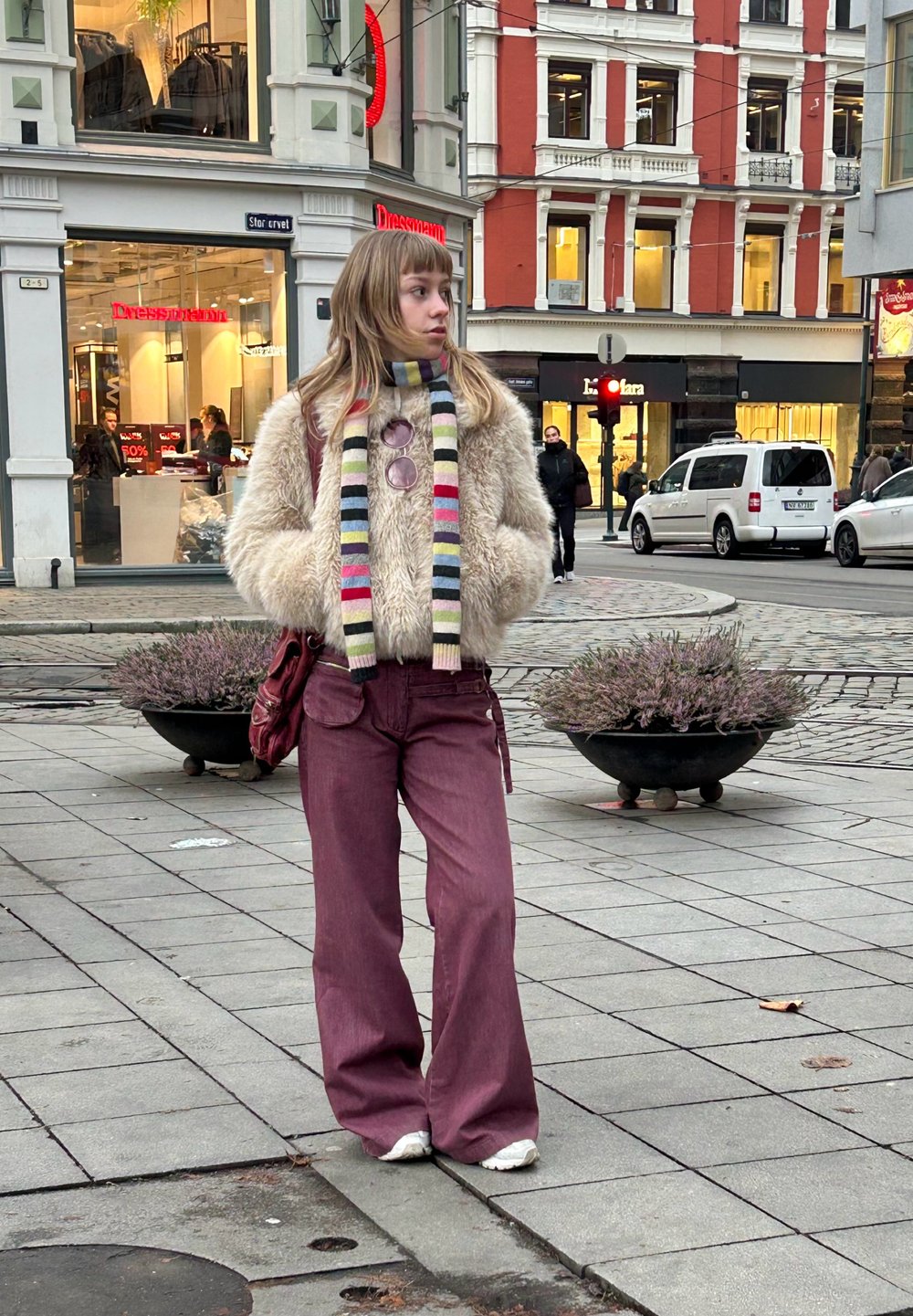 Jachetă crem pufoasă, eșarfă cu dungi multicolore, ochelari de soare rotunzi, pantaloni largi bordo, adidași albi, stând pe asfalt gri.