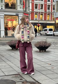 Jachetă crem pufoasă, eșarfă cu dungi multicolore, ochelari de soare rotunzi, pantaloni largi bordo, adidași albi, stând pe asfalt gri.