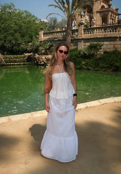 Top blanco sin mangas con un dobladillo con volantes, combinado con una falda larga blanca con sutiles lunares. Bolsa marrón, gafas de sol negras y palmeras de fondo.
