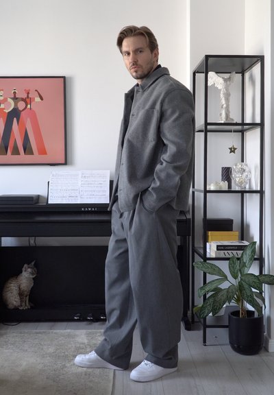 Chaqueta de lana gris con cuello, combinada con pantalones anchos grises. Zapatillas blancas. El fondo presenta un piano, un gato y estanterías decorativas.