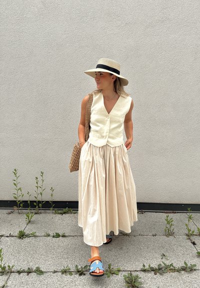 Un chaleco de lino color beige claro con escote en V, combinado con una falda larga y fluida color crema y sandalias azules. Una bolsa de mimbre completa el conjunto.