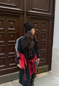 Trench-coat noir avec trois bandes blanches, bonnet noir, chemise à carreaux rouges nouée à la taille, sac à main rouge et pantalon camouflage devant une porte marron.