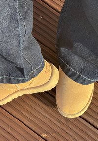 Pieds chaussés de bottes UGG en daim beige et pantalon foncé, debout sur une terrasse en bois avec des rainures et des vis visibles.