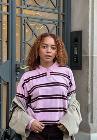 Młoda kobieta z kręconymi włosami, ubrana w różową koszulę w paski i beżowy płaszcz, stoi obok metalowej bramy i systemu wejściowego z klawiaturą.