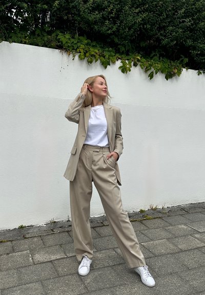 Blazer beige ajustado sobre una camiseta blanca, combinado con pantalones beiges holgados y zapatillas blancas. De pie contra una pared blanca.