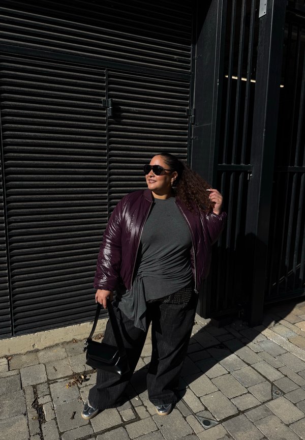 Una mujer con gafas de sol y una chaqueta de color ciruela está de pie en una acera de la ciudad, sostiene un bolso negro y se arregla la coleta.