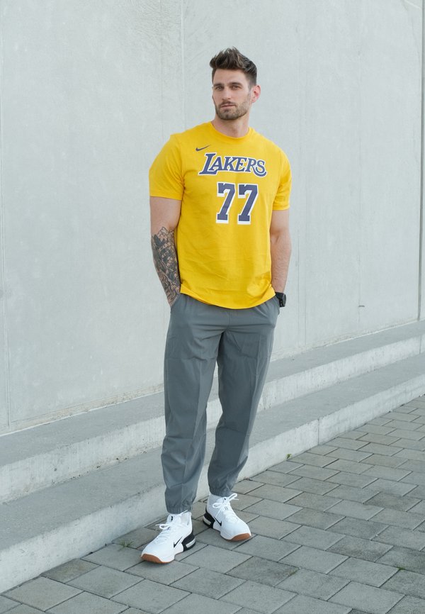 Man met tatoeage draagt een geel Lakers 77-shirt, grijze broek en witte sneakers, staand op een geplaveide stoep bij een betonnen muur.