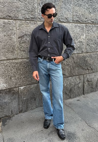 Camisa negra de manga larga con botones, jeans rectos de mezclilla azules claros, cinturón negro y zapatos negros pulidos sobre un fondo de piedra texturizada.