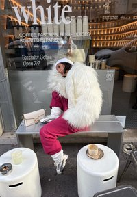 Personne en veste blanche duveteuse et pantalon rose assise sur un banc à l'extérieur d'un café, avec une tasse de café et un jus sur des tabourets à proximité.