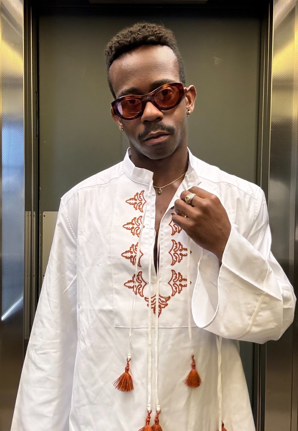 White shirt with red embroidery, loose fit, and long sleeves. Features decorative tassels and a placket design. Brown sunglasses.