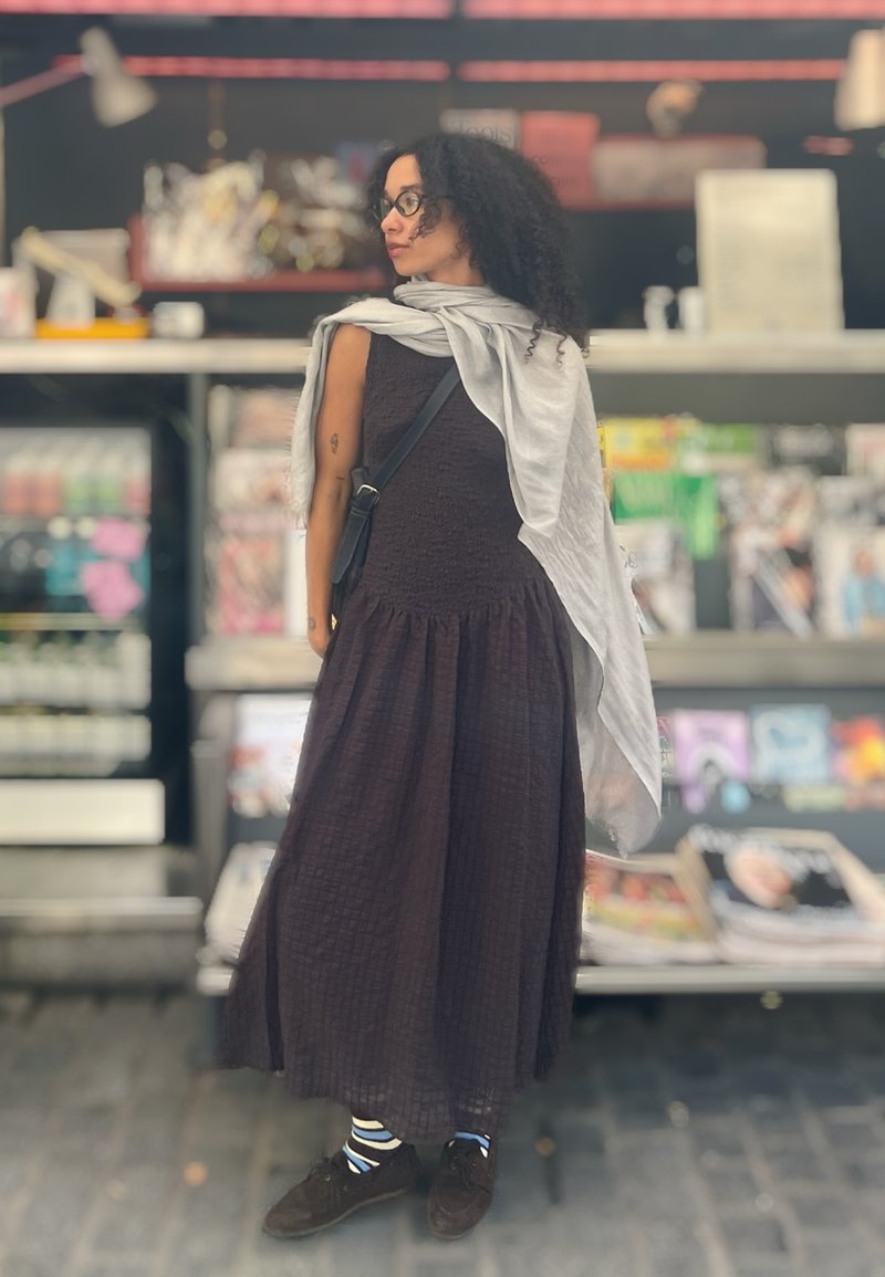 Mujer con cabello rizado que lleva gafas, un vestido largo oscuro, calcetines a rayas, zapatos oscuros y una bufanda gris clara, de pie junto a un quiosco de revistas.