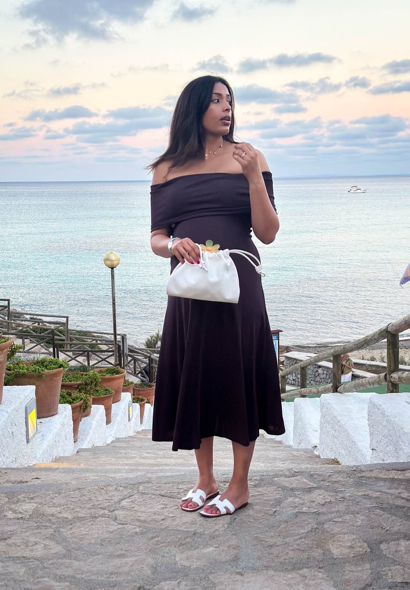 Vestido castanho escuro, com decote off-the-shoulder e silhueta fluida, combinado com uma mala branca de cordão e sandálias brancas, em degraus de pedra.